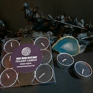 May include: A set of black tealight candles in a clear plastic container, with three loose candles. The label reads "Dark Moon Merchant Tealight Candles" with a website address. A blue and white geode slice and black floral accents are in the background.