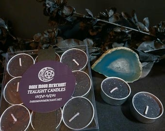 May include: A set of black tealight candles in a clear plastic container, with three loose candles. The label reads "Dark Moon Merchant Tealight Candles" with a website address. A blue and white geode slice and black floral accents are in the background.