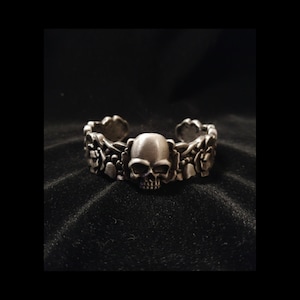 May include: A silver-toned cuff bracelet featuring a central skull design. The bracelet is adorned with floral and skull motifs along its band. The open-ended design allows for easy wear. The bracelet is displayed against a dark background.
