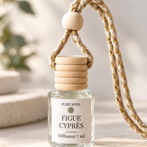 May include: A 7 ml glass car air freshener bottle with a light wood cap and a beige rope featuring gold metallic threading. The white label displays the text PURE SOYA, a small floral icon, FIGUE CYPRÈS, and the volume measurement of 7 ml.