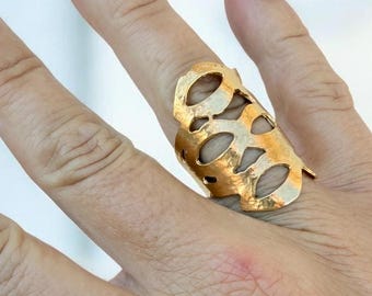 May include: A gold-colored statement ring with an open, cut-out design is displayed on a hand with long, burgundy-colored nails. The ring has an elongated shape with several oval cut-outs. The background is blurred, focusing attention on the jewelry.