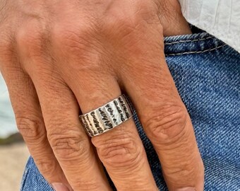 May include: A silver ring and bracelet with textured, horizontal lines. The ring is on a finger, and the bracelet is on the wrist. The jewelry is worn with a white shirt and blue jeans. The background is a beach scene.
