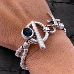 May include: A silver bracelet with a toggle clasp and a blue gemstone. The bracelet has a double strand of small, round beads. A silver ring with a unique design is also visible.