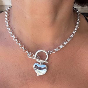 May include: A silver-toned necklace with a heart pendant. The necklace features a chunky chain and a toggle clasp. The heart pendant is smooth and reflective, adding a touch of elegance to the jewelry.