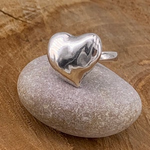 May include: A silver heart-shaped ring with a smooth, polished finish. The ring is displayed on a light gray stone.