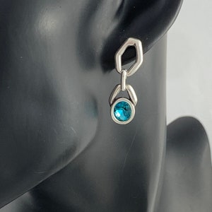 May include: Silver-toned dangle earring with a geometric design. The earring features a small, round, bright blue gemstone at the bottom. The top portion has a unique hexagonal shape. The earring is displayed on a black mannequin head.