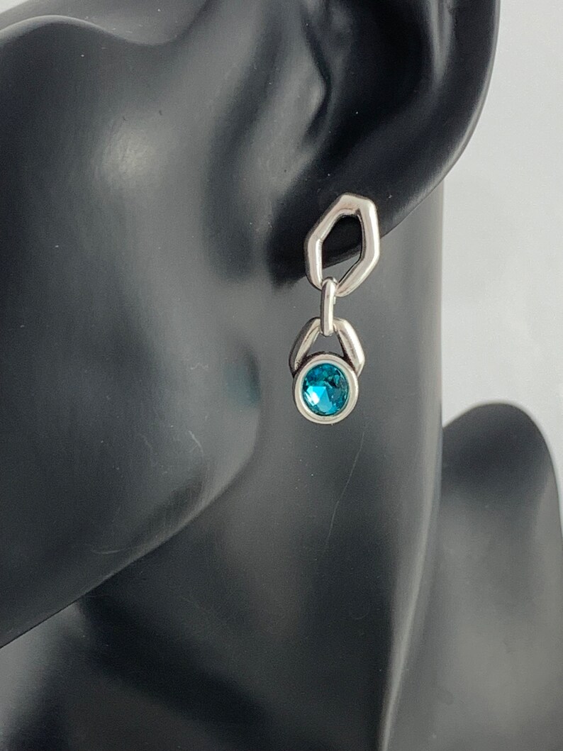 May include: Silver-toned dangle earring with a geometric design. The earring features a small, round, bright blue gemstone at the bottom. The top portion has a unique hexagonal shape. The earring is displayed on a black mannequin head.