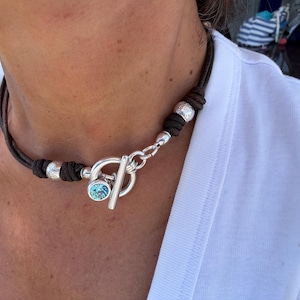 May include: A brown leather necklace with silver accents. The necklace features a toggle clasp and a light blue gemstone. The necklace is worn over a white shirt.
