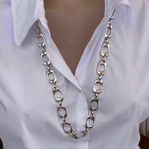 May include: A silver chain necklace with large, oval-shaped links. The chain is worn on a white shirt.
