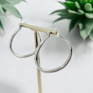 Puede incluir: Un par de pendientes de aro plateados con forma de l&aacute;grima, expuestos en un soporte dorado. Los pendientes tienen un acabado pulido. El fondo incluye plantas verdes y una superficie blanca.