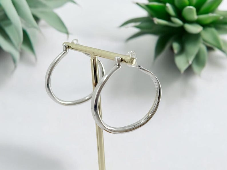 Puede incluir: Un par de pendientes de aro plateados con forma de l&aacute;grima, expuestos en un soporte dorado. Los pendientes tienen un acabado pulido. El fondo incluye plantas verdes y una superficie blanca.