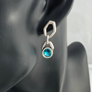 May include: A silver earring with a geometric design. The earring features a small, round, bright blue gemstone set in a silver bezel. The earring is attached to a silver, open-sided hexagon shape.