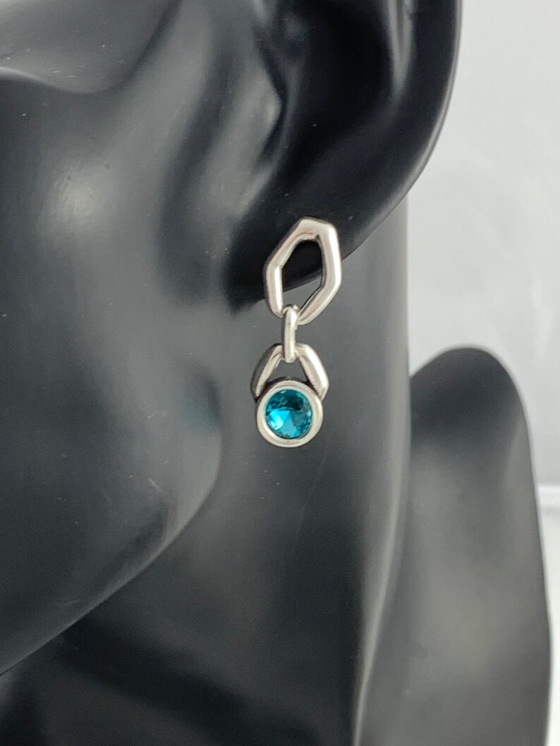 May include: A silver earring with a geometric design. The earring features a small, round, bright blue gemstone set in a silver bezel. The earring is attached to a silver, open-sided hexagon shape.