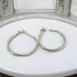 Puede incluir: Un par de pendientes de aro plateados. Los pendientes tienen un aspecto texturizado y martillado y se muestran sobre una superficie blanca. Los pendientes son redondos y tienen cierre de poste.