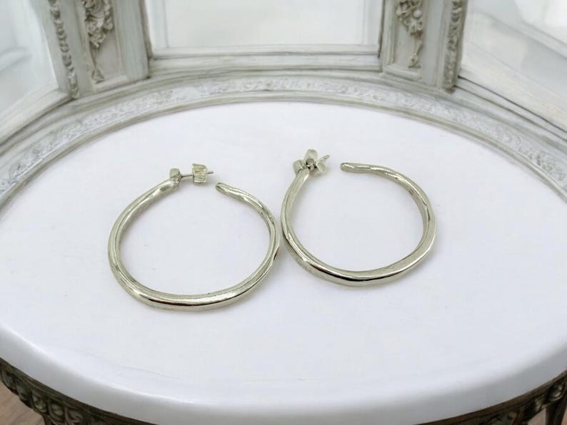 Puede incluir: Un par de pendientes de aro plateados. Los pendientes tienen un aspecto texturizado y martillado y se muestran sobre una superficie blanca. Los pendientes son redondos y tienen cierre de poste.