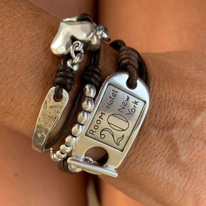 Könnte beinhalten: Mehrsträngiges Armband mit einem silbernen Herzanhänger, einem strukturierten Silberstab und einem Anhänger mit der Aufschrift "Room Hotel 20 New York". Das Armband enthält braune Lederkordeln und eine Reihe von Silberperlen. Das Armband wird am Handgelenk getragen.