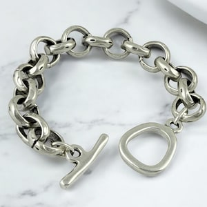 May include: Silver-toned bracelet featuring a chunky chain design with a toggle clasp. The bracelet has a series of interconnected, rounded links. The clasp includes a T-bar and a triangular loop for secure fastening. The jewelry is displayed on a white surface.