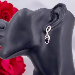 May include: Silver-toned drop earrings with a black oval stone. The earrings are displayed on a black mannequin head, with red roses in the background. The earrings have a geometric design.