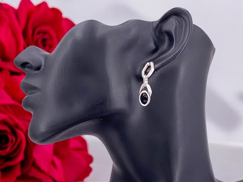 May include: Silver-toned drop earrings with a black oval stone. The earrings are displayed on a black mannequin head, with red roses in the background. The earrings have a geometric design.