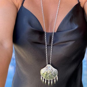 Puede incluir: Collar con colgante plateado con un diseño de concha texturizada y elementos colgantes en forma de cono. El collar de cadena larga se lleva sobre una blusa de satén negro.