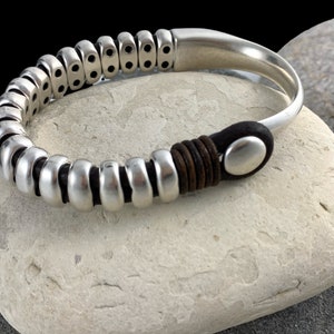 Puede incluir: Brazalete de puño plateado con un diseño único. El brazalete presenta una serie de segmentos metálicos redondeados y conectados en un lado, que se transforman en una banda lisa y curvada. El cierre está acentuado con cuero marrón y una envoltura de cuero negro.