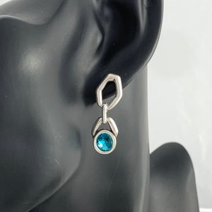 May include: A silver earring with a geometric design. The earring features a hexagonal top, a connecting link, and a round, blue gemstone. The earring is displayed on a black mannequin head.