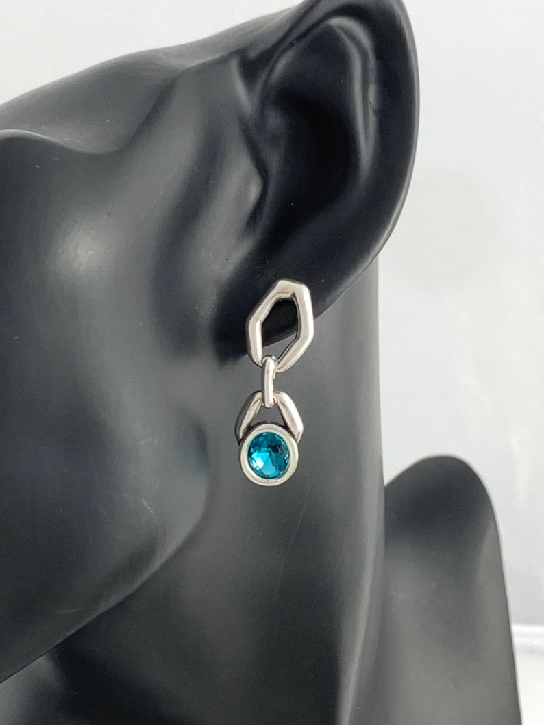 May include: A silver earring with a geometric design. The earring features a hexagonal top, a connecting link, and a round, blue gemstone. The earring is displayed on a black mannequin head.