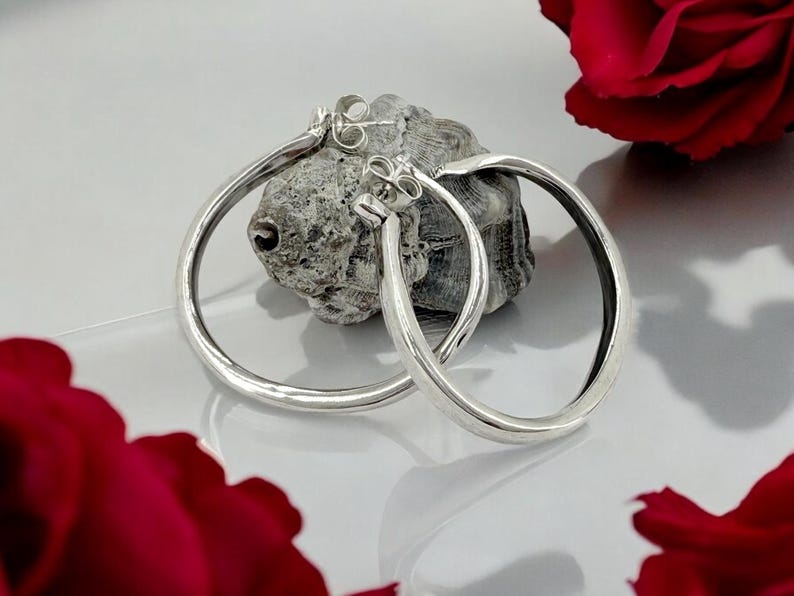 May include: A pair of silver hoop earrings with a hammered texture, resting on a textured gray stone. The earrings are round and shiny, reflecting light. Red rose petals are in the foreground.
