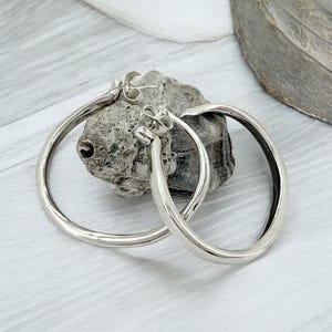 Puede incluir: Un par de pendientes de aro plateados con un acabado liso y pulido. Los pendientes son circulares y tienen un dise&ntilde;o simple y elegante. Se exhiben sobre una piedra gris texturizada contra un fondo gris claro y blanco.