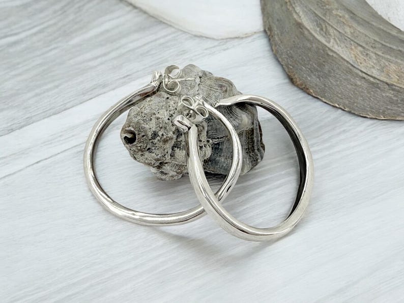 Puede incluir: Un par de pendientes de aro plateados con un acabado liso y pulido. Los pendientes son circulares y tienen un dise&ntilde;o simple y elegante. Se exhiben sobre una piedra gris texturizada contra un fondo gris claro y blanco.