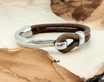May include: A brown leather bracelet with a silver metal clasp and a knot design. The bracelet is on a wooden surface.
