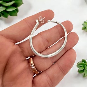 Puede incluir: Un pendiente de aro plateado con un dise&ntilde;o liso y redondeado. El pendiente se sostiene en una mano, mostrando su superficie pulida. Una pieza de joyer&iacute;a cl&aacute;sica, adecuada para el uso diario.