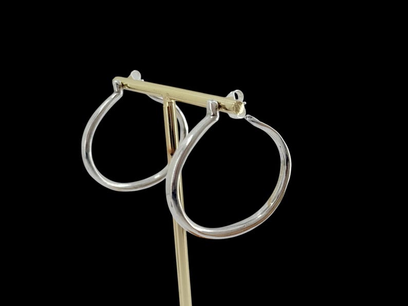 Puede incluir: Un par de pendientes de aro plateados con forma de l&aacute;grima, expuestos sobre un soporte dorado para pendientes sobre un fondo negro. Los pendientes tienen un acabado pulido y reflejan la luz.