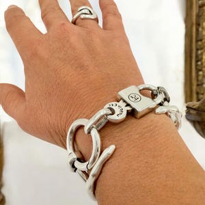 Könnte beinhalten: Ein silbernes Armband mit einem Schlossverschluss. Das Schloss hat die Nummer "24" eingraviert. Das Armband besteht aus dicken, massiven Gliedern.