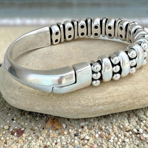 Puede incluir: Un brazalete plateado con un diseño único. Presenta una combinación de secciones lisas y curvas y un patrón segmentado y texturizado. El cierre es sencillo y seguro. El brazalete se muestra sobre una piedra clara.