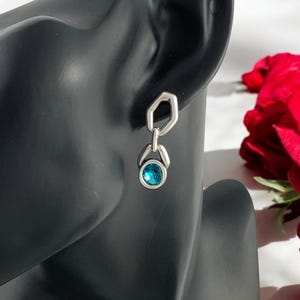 May include: A silver earring with a geometric design. The earring features a hexagonal top connected to a teardrop-shaped setting holding a round, bright blue gemstone. The earring is displayed on a black mannequin head.