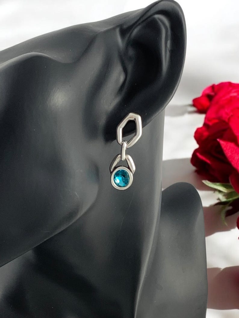 May include: A silver earring with a geometric design. The earring features a hexagonal top connected to a teardrop-shaped setting holding a round, bright blue gemstone. The earring is displayed on a black mannequin head.