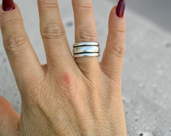 May include: A silver ring with a thick band and three stacked layers is worn on a finger. The hand has long nails painted a deep burgundy color. The background is a light gray.