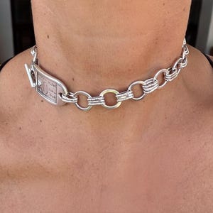 Può includere: Collana a catena color argento con un ciondolo rettangolare. Il ciondolo presenta un testo inciso ed è collegato a una serie di grandi segmenti di catena collegati. La collana è indossata intorno al collo.