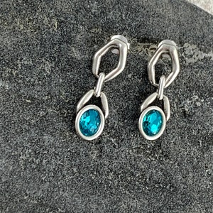 May include: A pair of silver-toned earrings with a unique design. Each earring features a small, open geometric shape connected to an oval-shaped setting holding a vibrant, teal-colored gemstone. The earrings are displayed on a textured, dark gray surface.