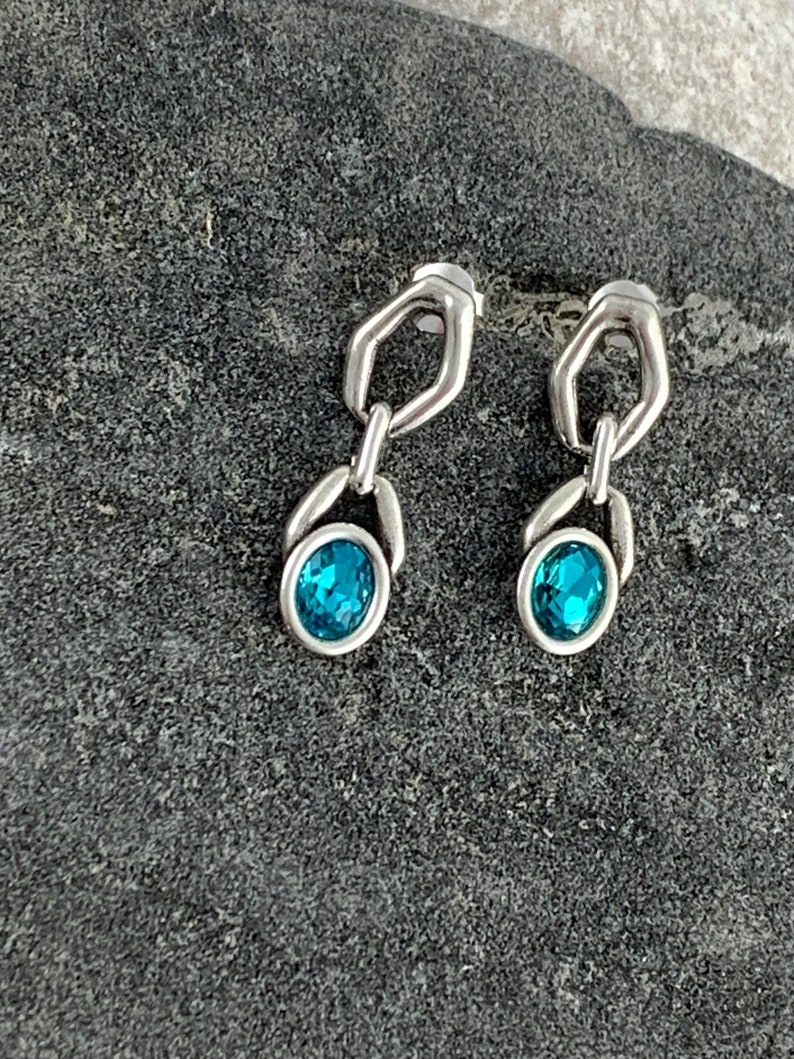 May include: A pair of silver-toned earrings with a unique design. Each earring features a small, open geometric shape connected to an oval-shaped setting holding a vibrant, teal-colored gemstone. The earrings are displayed on a textured, dark gray surface.