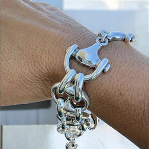 May include: A silver bracelet with a distinctive design, featuring interlocking links and a decorative clasp. The bracelet is worn on a wrist, highlighting its intricate details and polished finish. The jewellery piece has a modern, eye-catching aesthetic.