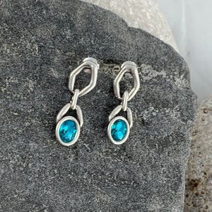 May include: A pair of silver-toned earrings with a geometric design. Each earring features a blue oval-shaped gemstone. The earrings are displayed on a dark gray stone surface.