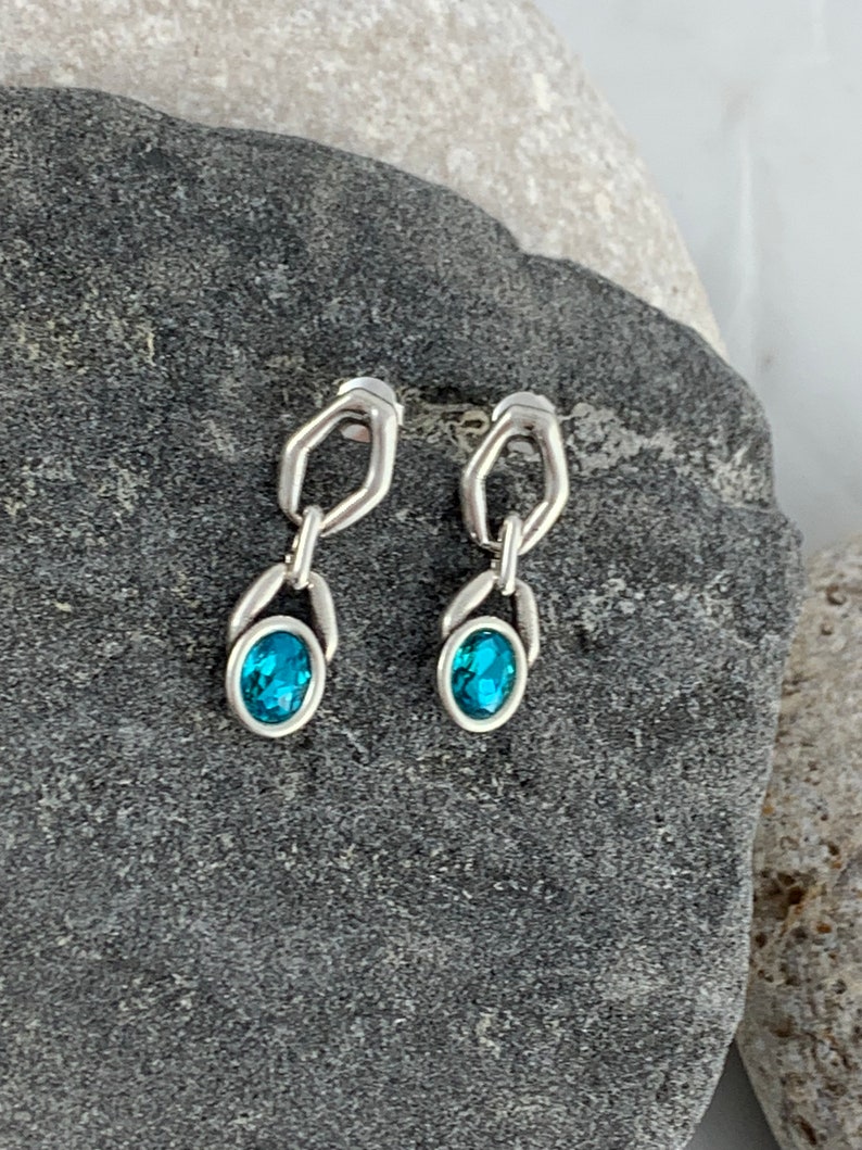 May include: A pair of silver-toned earrings with a geometric design. Each earring features a blue oval-shaped gemstone. The earrings are displayed on a dark gray stone surface.