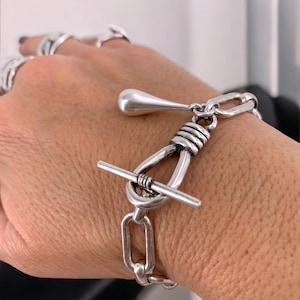 May include: A silver bracelet with a toggle clasp. The bracelet features a chain link design with a teardrop-shaped charm and a bar closure. The bracelet is worn on a wrist, with other silver rings visible in the background.