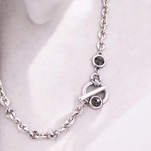 May include: A silver-toned necklace with a chain link design. The necklace features a toggle clasp with a circular element and a bar closure. Two small, dark gray gemstones are set into the necklace.