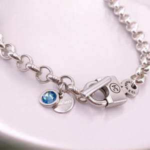 Può includere: Un braccialetto a catena d'argento con un ciondolo di pietra blu e una chiusura a lucchetto. Il lucchetto ha il numero "24" inciso. La catena è di stile curb.