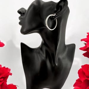 Puede incluir: Pendientes de aro plateados exhibidos en una cabeza de maniqu&iacute; negra. Los pendientes son circulares y tienen un acabado pulido. Rosas rojas visibles en el fondo, sobre un fondo blanco.