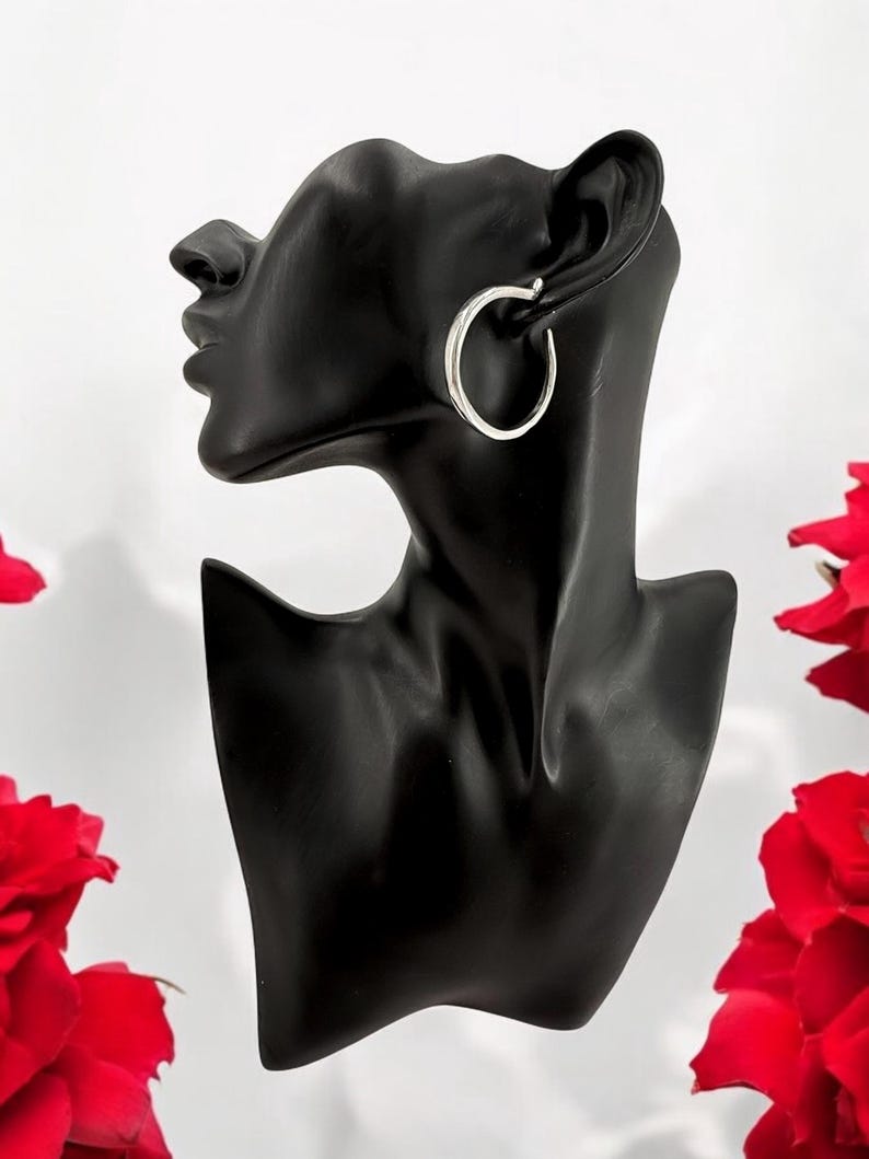 Puede incluir: Pendientes de aro plateados exhibidos en una cabeza de maniqu&iacute; negra. Los pendientes son circulares y tienen un acabado pulido. Rosas rojas visibles en el fondo, sobre un fondo blanco.