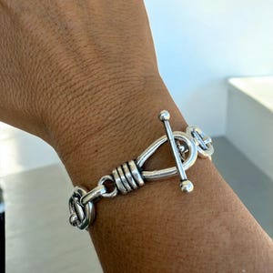 May include: A silver bracelet with a toggle clasp. The bracelet has interconnected links and a central design with wrapped detailing. The toggle clasp is a bar that fits through a loop. The jewellery is worn on a wrist.